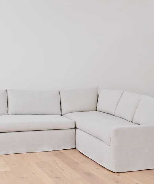The Harbor Sectional in light gray Belgian flax linen features clean lines and rests in a corner with white walls and light wood flooring, capturing the relaxed elegance of Jenni Kayne Furniture.