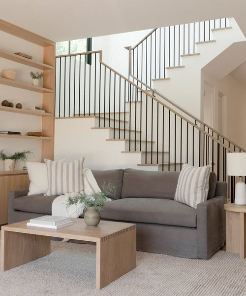 A modern living room with the Harbor Sofa in charcoal linen, striped pillows, a light wood coffee table, built-in shelves, and a staircase with black railings; neutral tones and minimalist décor create a calm, airy feel.