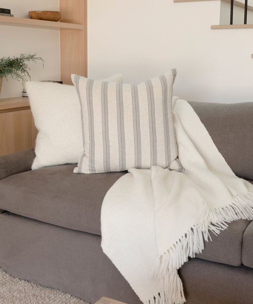 A handmade Harbor Sofa in charcoal linen, crafted in Los Angeles, is styled with a white fringed throw over the armrest, two accent pillows—one plain white and one with gray stripes—and a wooden shelf with a plant in the background.