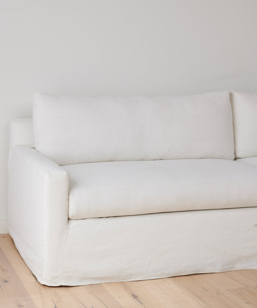 The Harbor Sofa, featuring ivory linen and plush cushions, stands on a light wooden floor against a plain white wall. This modern, minimalist piece is handmade in Los Angeles.
