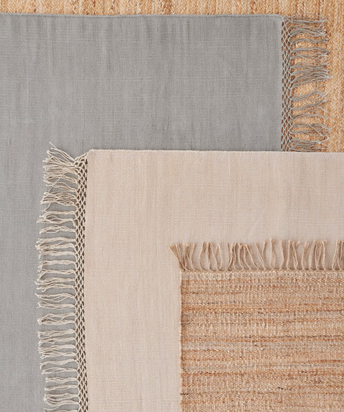 Three rectangular woven rugs are layered: a gray rug on the left, a beige handwoven rug in the center, and the Hudson Jute Rug with coarse texture underneath.