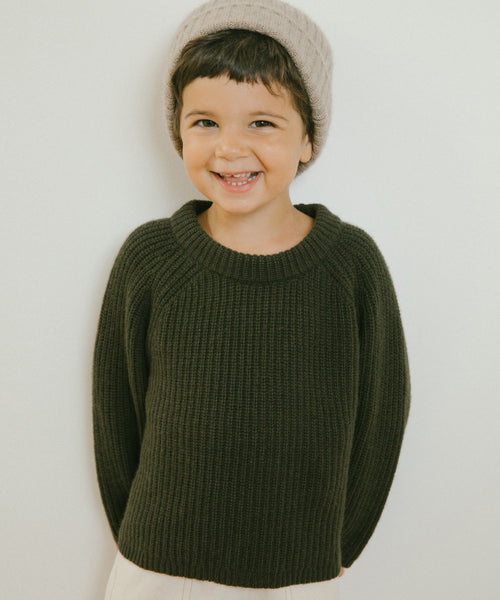A young child smiles at the camera in a Kids Fisherman Sweater (olive) and a light gray beanie, standing against a plain white background.