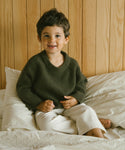 A young child with short brown hair, smiling and wearing an olive Kids Fisherman Sweater and light pants, sits on a bed with white sheets against a wooden wall.