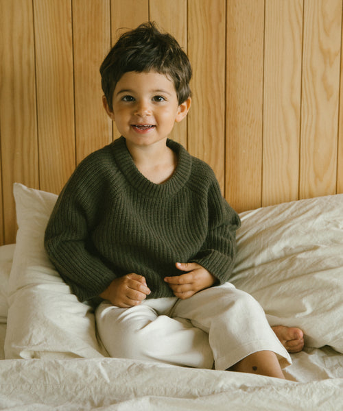 A young child with short brown hair, smiling and wearing an olive Kids Fisherman Sweater and light pants, sits on a bed with white sheets against a wooden wall.