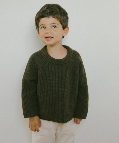 A young child with short brown hair wears the Kids Fisherman Sweater in dark green and light pants, standing against a plain light background and smiling slightly while looking to the side.