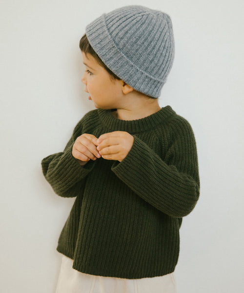 A young child in an olive Kids Fisherman Sweater and a gray knit beanie looks to the side against a plain light background.
