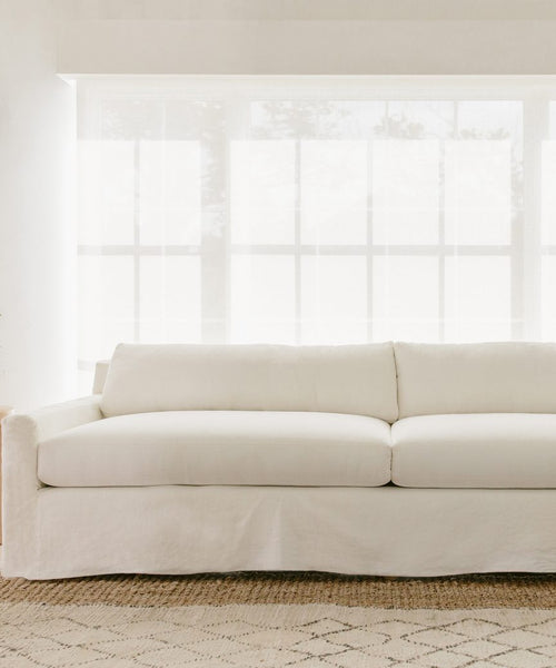 A bright, minimalist room features a modern white sofa upholstered in Fabric Swatch Ivory Linen, positioned before large windows with sheer curtains and set atop a light patterned rug.