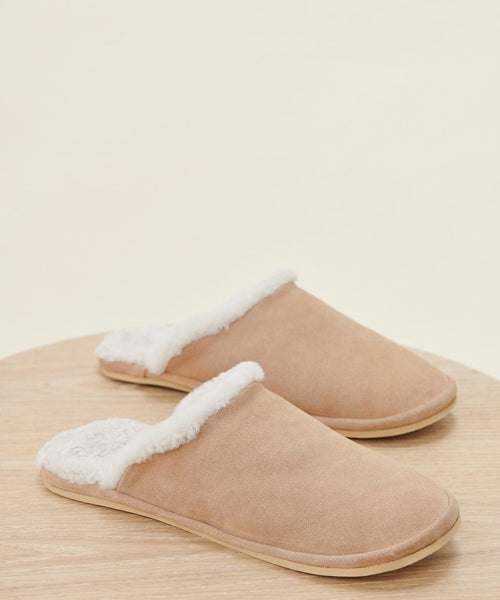 The Lodge Outdoor Slipper features a tan suede upper and white shearling lining, shown on a light wooden surface against a neutral background.