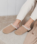 Someone wearing Lodge Outdoor Slippers, featuring a beige suede upper and cozy shearling lining, pairs them with light sweatpants while sitting on a textured beige couch with a matching rug. Only their lower legs and feet are visible.