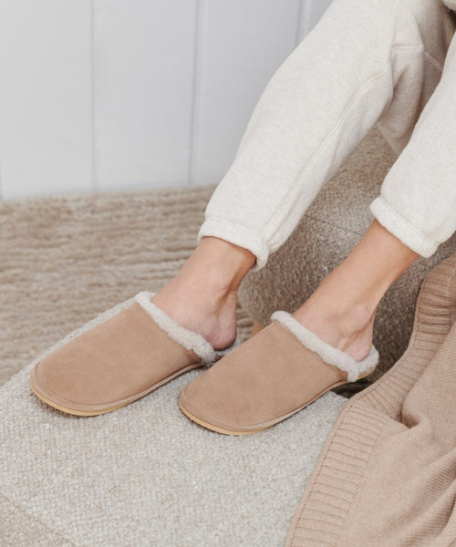 Someone wearing Lodge Outdoor Slippers, featuring a beige suede upper and cozy shearling lining, pairs them with light sweatpants while sitting on a textured beige couch with a matching rug. Only their lower legs and feet are visible.