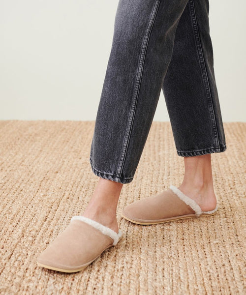 A person wearing cropped black jeans and the Lodge Outdoor Slipper in beige with cozy shearling lining, standing on a woven carpet.