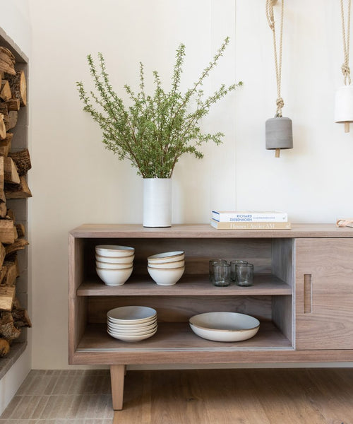 A Mandeville Credenza holds stacked plates, bowls, and glasses. A tall white vase with green branches sits on top beside a small stack of books. Firewood is stacked to the left and hanging lights glow to the right.