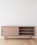 The Mandeville Credenza features a minimalist walnut design with angled legs, a left sliding door, and three open right shelves. Its shown against a plain white wall on a light wooden floor.