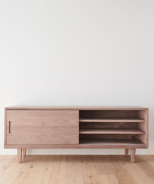 The Mandeville Credenza features a minimalist walnut design with angled legs, a left sliding door, and three open right shelves. Its shown against a plain white wall on a light wooden floor.