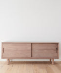 The Mandeville Credenza stands against a plain white wall on a light wood floor, featuring minimalist design with sliding doors and angled legs.