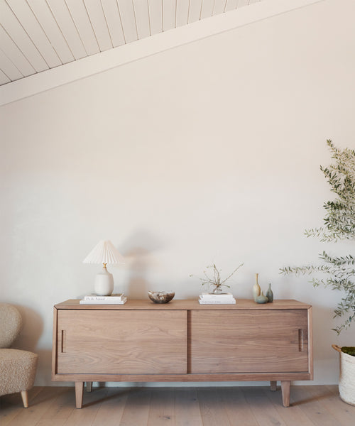 The Mandeville Credenza showcases a lamp, books, bowl, vases, and a small plant against a light wall in a modern space with a neutral palette.