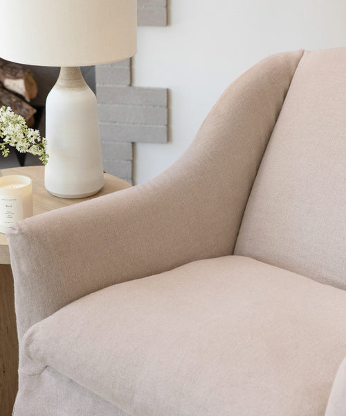 A close-up of the Miramar Chair, upholstered in beige Belgian linen and handmade in the USA, sits by a round wooden side table displaying a white ceramic lamp, small candle, and a vase of white flowers.