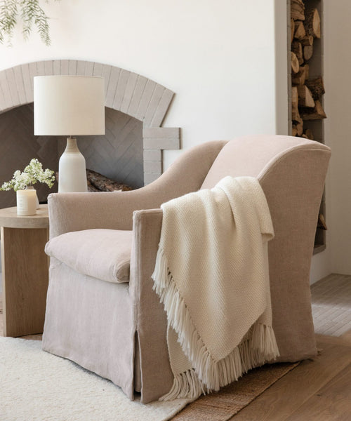A Miramar Chair upholstered in beige Belgian linen, adorned with a white fringed throw on one arm, is placed beside a wooden side table holding a lamp and flowers, positioned before a fireplace stacked with wood.