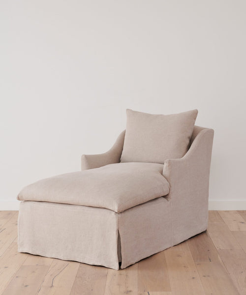 A Miramar Chaise in beige Belgian linen, complete with cushion and pillow, is placed on a light wood floor against a white wall.