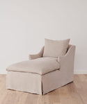 The Miramar Chaise Slipcover in Belgian linen covers a modern beige upholstered chaise lounge, set on a light wooden floor against a plain white wall.