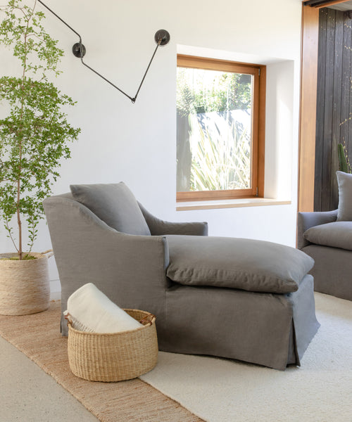 A modern living room featuring the Miramar Chaise, a large gray armchair, a wicker basket with a white blanket, a potted plant, woven rug, and ample natural light from the window.