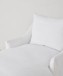 A close-up of a white upholstered armchair featuring the Miramar Chaise Slipcover in Belgian linen and a soft cushion, set against a plain white wall for a cozy, minimalist look.