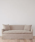 The Miramar Sofa Slipcover, in minimalist beige Belgian linen with two matching cushions, sits against a plain white wall on a light wooden floor.