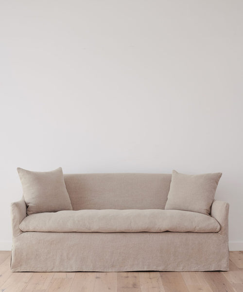 The Miramar Sofa Slipcover, in minimalist beige Belgian linen with two matching cushions, sits against a plain white wall on a light wooden floor.