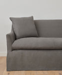 The Miramar Sofa, upholstered in gray Belgian linen with feather down fill and a matching cushion, sits against a plain white wall on a light wooden floor. Only the left side of the sofa is visible.