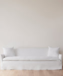 The Miramar Sofa Slipcover, in minimalist white linen with two matching pillows, sits against a plain light-colored wall on a light wood floor.