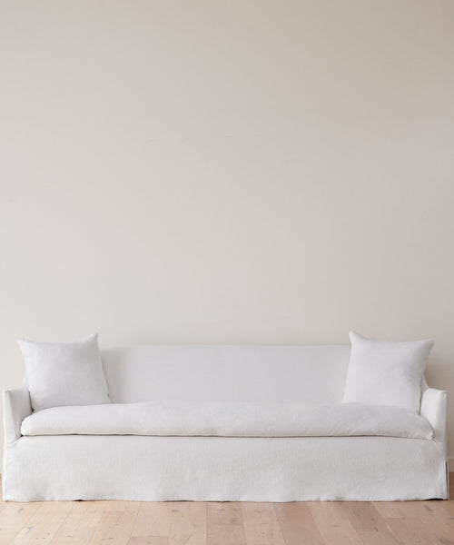 The Miramar Sofa Slipcover, in minimalist white linen with two matching pillows, sits against a plain light-colored wall on a light wood floor.