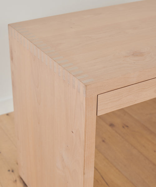 A close-up of the Newport Desk highlights its smooth solid wood surface, visible wood grain, and precise dovetail joinery at the corner, set on a light-colored wooden floor.