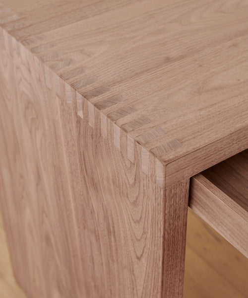 Close-up of a Newport Desk corner showcases precise dovetail joinery and rich walnut wood grain, emphasizing fine craftsmanship. The smooth, unfinished surface highlights the natural beauty of the material.