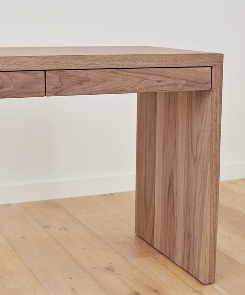 The Newport Desk features a minimalist walnut design with clean lines and two slim drawers, shown against a plain white wall atop a light wood floor.
