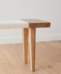 A close-up of the Oak Day Bench, featuring angled legs and a woven white seat, placed on a light wood floor against a plain white wall.