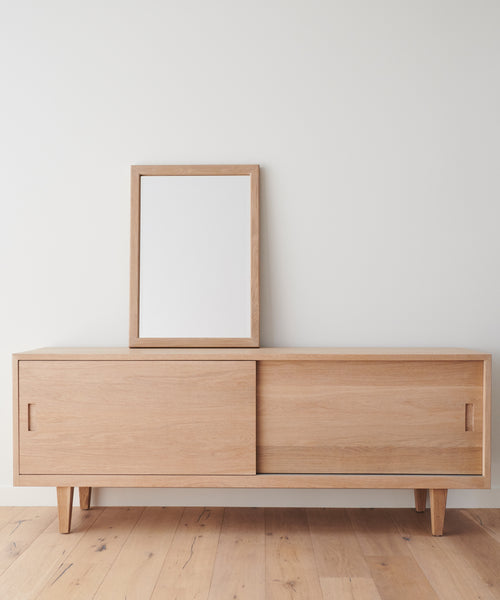 A light wooden sideboard with sliding doors stands against a white wall. An Oak Wall Mirror rests on top, reflecting the plain wall. The floor is light wood, matching the sideboard.
