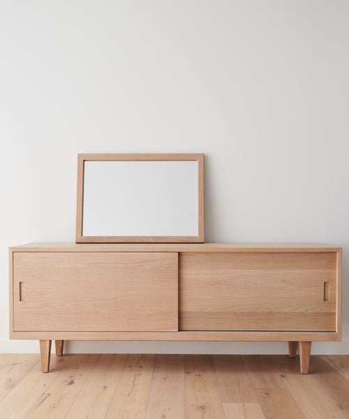 A light wood sideboard with sliding doors stands against a white wall. An Oak Wall Mirror with a solid wood frame rests slightly off-center on top. The light wood floor enhances the minimalist, modern look.