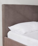 A close-up of the Pacific Bed, featuring a tall, upholstered headboard in gray Belgian linen with a white pillow and crisp white bed sheets, set against a plain light-colored wall.