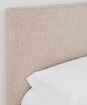 A close-up of the Pacific Bed’s textured Belgian linen upholstered headboard sits beside a neatly made bed with white linens and a white pillow, set against a plain white wall.