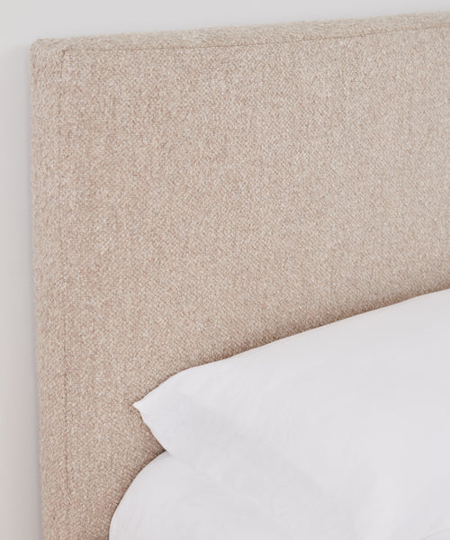 A close-up of the Pacific Bed’s textured Belgian linen upholstered headboard sits beside a neatly made bed with white linens and a white pillow, set against a plain white wall.