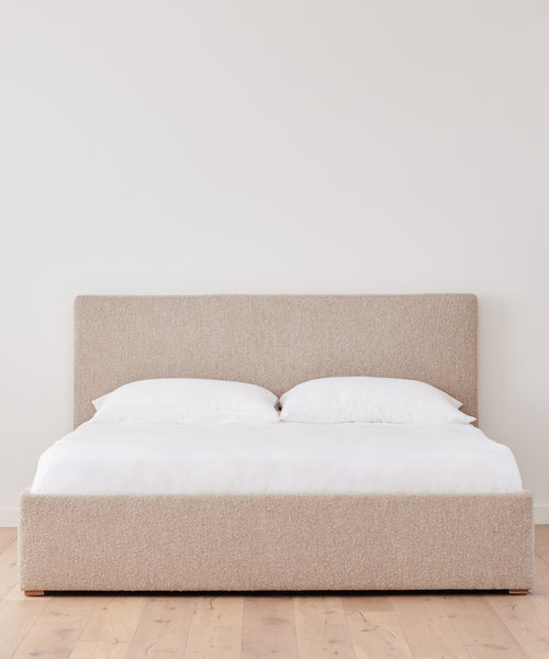 The Pacific Bed, featuring modern beige upholstery and a tall Belgian linen headboard, is styled with white bedding and two pillows in a minimal room with light wood floors and a plain white wall.