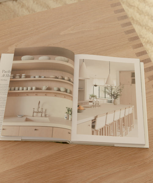 An open copy of Pacific Natural At Home rests on a wooden table, featuring photos of a minimalist kitchen with light wood cabinets, open shelves, a long counter, and white bar stools—highlighting California style.