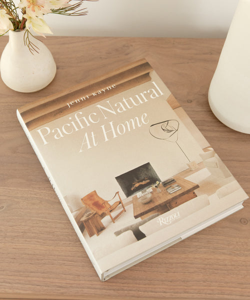 The book Pacific Natural At Home rests on a wooden table near a white vase with flowers and a round white container. Its cover shows a minimalist living room scene inspired by California style.