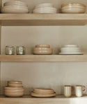 Three wooden shelves display neatly stacked Pacific Salad Plates in neutral tones, stoneware dinner plates, mugs, and two short glass tumblers, all arranged against a light-colored wall.