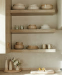 Three wooden shelves display neatly stacked neutral-toned plates, bowls, and mugs, including the Pacific Serving Bowl. Below, a counter holds oil dispensers, a white flower vase, cutting board, and sliced lemons in this minimalistic kitchen.
