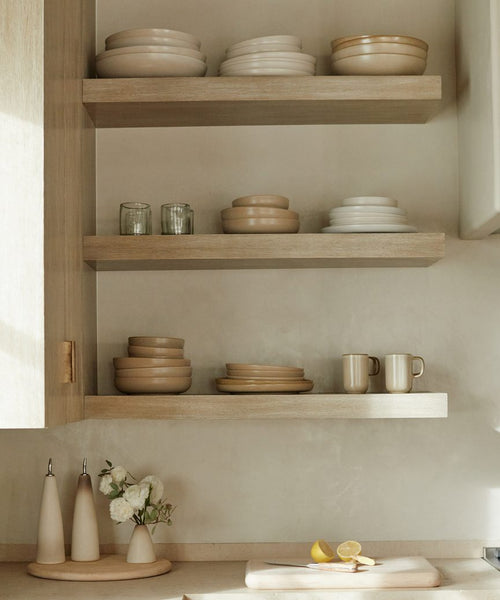 Three wooden shelves display neatly stacked neutral-toned plates, bowls, and mugs, including the Pacific Serving Bowl. Below, a counter holds oil dispensers, a white flower vase, cutting board, and sliced lemons in this minimalistic kitchen.