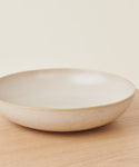 The Pacific Serving Bowl, a shallow and round beige stoneware piece, is placed on a light wooden surface with a plain off-white background.
