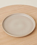 A plain, round, beige Pacific Platter stoneware serving plate rests on a light wooden surface, photographed from above.