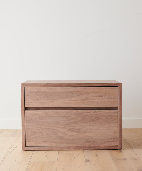 The Palms Nightstand, a minimalist light wood piece with two drawers, sits against a white wall on a light wooden floor, pairing perfectly with solid walnut furniture for a beautifully cohesive look.