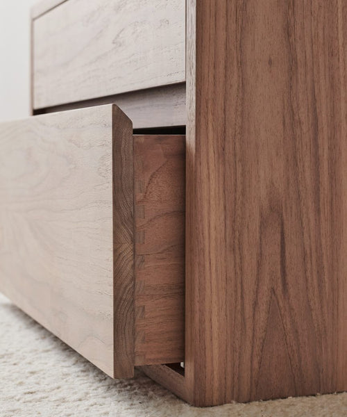 Close-up view of the Palms Nightstand, crafted from solid walnut. One drawer is partially open, showing precise dovetail joints. This elegant nightstand rests on a light-colored textured carpet.